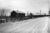 Ett stort bostadshus med tillhörande uthus ligger innanför en gärdesgård. På vägen utanför är det snö och snövallar vid sidorna.