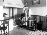 En äldre kvinna sitter vid sin spinnrock. Till vänster är eldstaden med en järnspis och till höger står en kökssoffa av trä där det ligger färdigspunna garnhärvor.