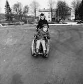 Ett barn kör ett annat barn i rullstol på grusplanen öster om Stora Skolan vid Stretereds skolhem, cirka 1959-1980. Förskolan syns i bakgrunden.