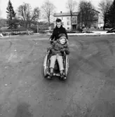 Ett barn kör ett annat barn i rullstol på grusplanen öster om Stora Skolan vid Stretereds skolhem, cirka 1959-1980. Förskolan syns i bakgrunden.
