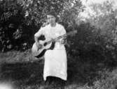 Helga Börjesson, född Lidman, sitter utomhus och spelar gitarr i Kållered cirka 1930. Familjen Börjesson var sommargäster i Hallen. Familjen bodde annars på Lilla Vegagatan i Göteborg.