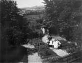 Utsikt från balkongen på Kvarnbygatan 23, 1920-tal. I trädgården syns några av husets invånare: Axel och Olivia Andersson med sina barn Arne, Inga och Astrid. 
Sonen Arne tog sig senare namnet Lundälv. 
Utsikt över de platser där Nya torget och Bosgården sedan fanns.