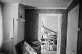 Bostadshus Roten M 11-12, okänt årtal. Ett rum med en tapetserad vägg, cykel och diverse prylar.