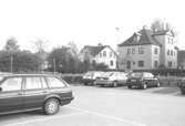 Parkeringsplats med villor i bakgrunden på Barnhemsgatan i Broslätt. Bilden är tagen under 1990-talet.