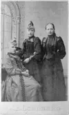 Tre kvinnor uppställda i en fotoateljé år 1900. Amalia Precht-Reuter (1871-1956) står i mitten. Hon var dotter till sockerbruksmästaren Precht-Reuter vid Korndals bruk, senare anställd vid Carnegies bruk.
