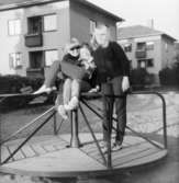 Tre flickor (Eija, Ann-Charlotte och Annette) står på en karusell i en lekpark. Brunnsgatan 4 och 6 syns i bakgrunden. 1960-tal.