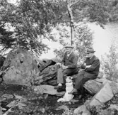 Albert Persson och Sigurd Olsson sittandes vid Skärgubbens ula (grotta) på Lillön i Nordsjön vi Långö, 1959. 
Sigurd Olsson var högt uppsatt kommunalman i Lindomes kommun.
Hunden hette Laika efter rymdhunden, ägdes av Albert P.