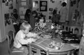 Två barn sitter och pysslar vid ett bord. I bakgrunden syns barn och en vuxen som tittar på uppsatta saker på en vägg. Utställningsvernissage av och om Katrinebergs daghem på Mölndals museum 1993-09-10.