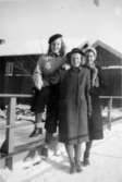 Astrid Karlssons (gift Garthman) kompisar sitter/står vid en bro i ett vinterklätt Kvarnbyn. Karin Karlsson står i mitten, de två andra är okända för givaren. 1930-tal.
