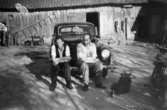 Birger och Herbert Lyckö sitter på stänkskärmen till en Opel Kapitän. På marken vid sidan om ligger en hund. I bakgrunden ser man en lada med en stege hängandes på väggen. Kvarnfallet i Mölndal, 1940-50-tal.