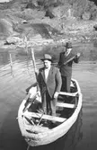 Från vänster: Helmer Garthman och Dimitri Korski står i en eka som ligger i vattnet. De är både fiske- och arbetskamrater. Klippor syns i bakgrunden vid Näset, 1960-tal.