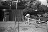 Främst i bild ser man en sandlåda och gungställning. Ett barn och en man står utanför. Mannen står böjd och sysslar med något. I bakgrunden syns ett förråd samt en bil. Katrinebergs daghem, 1992-93.