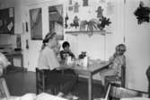 En kvinnlig förskollärare och tre pojkar sitter inomhus vid ett lågt fyrkantigt bord och fikar på Katrinebergs daghem. Ovanför dem på väggen sitter några uppklistrade teckningar samt en uppsatt hylla med växter på. Snett i bakgrunden till vänster syns ett par fönsterdörrar.