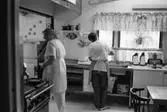 Christina Brussard och Kate Palm i köket på Katrinebergs daghem, 1992-93. Båda bär förklädde. Den ena kvinnan står vid sidan av spisen, den andra kvinnan diskar. På hennes t-shirt står 