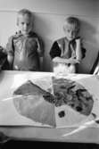 Två pojkar, iklädda förkläde, står uppställda bakom ett bord där det ligger en stor målad teckning, dekorerad med bl.a blad. Båda gossarna tittar mot kameran. Katrinebergs daghem, 1992-93.