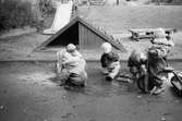 Fyra små barn leker utomhus i en vattenpöl. Ett barn sitter på en trehjuling. Samtliga är klädda i regnkläder och gummistövlar. I bakgrunden ser man en sandlåda med en liten rutschkana och längre bak ser man en större rutschkana på en kulle. Katrinebergs daghem, 1992.