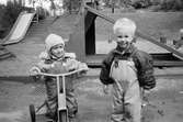 Två små pojkar, tittar leende mot fotografen. De är i lekplatsens sandlåda, iklädda regnkläder. Den högra pojken sitter på en trehjuling och den andre står bredvid. I bakgrunden ser man rutschkanor och klätterställning. Katrinebergs daghem, 1992.