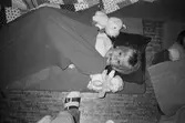 Ett barn ligger på en madrass placerad på golvet. Barnet tittar mot fotografen och har en filt över sig. På varje sida om barnet ligger ett gosedjur. Man skymtar foten och benet av läraren som har tagit bilden. Lille Skutt, Katrinebergs daghem, 1992.