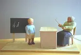 En skolpojke står framför en griffeltavla. Vid sidan av sitter skolfröken på en stol bakom sin kateder. Hon pekar med en pinne. Harry Bergmans 