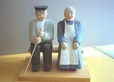 En äldre gubbe och gumma sitter bredvid varandra. Gubben har hatt och håller en käpp i handen. Gumman är klädd i vitt förkläde och har lagt sina händer i knät. Harry Bergmans 