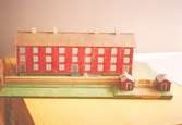 Ett rött bostadshus med tio fönster i rad, tre våningar högt, och tre entréer. På taket finns fyra skorstenar. Vid sidan av ligger två mindre stugor. Huset är en kopia av 