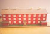 Ett rött bostadshus med tio fönster i rad, tre våningar högt, och tre entréer. På taket finns fyra skorstenar. Vid sidan av ligger två mindre stugor. Huset är en kopia av 