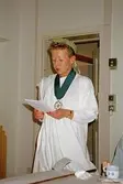 Pia Persson, iklädd toga, lagerkrans och medalj läser ur ett ark. Detta i samband med avtackning av receptionist Kim Linder på Mölndals museum 1994.