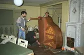 Tre män som monterar ihop en nyligen förflyttad flygel. Vid sidan om står en vacker kakelugn. Gunnebo slott, 1988.