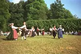 Ett folkdanslag som dansar omkring på Gunnebo slotts gräsmattor. Några sitter runt om och beskådar dansen.