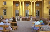 En orkester, bestående av sju män, spelar musik inför publik på Gunnebo slotts framsida. Publiken sitter på bänkar.