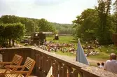 Människor sitter på gräsmattan och lyssnar till Lasse Berghagen. En tillfällig scen är uppbygd för evemenaget. Gunnebo slotts terrass, på baksidan.