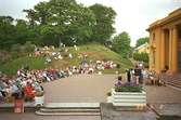 Människor som sitter och tittar på ett musik- och teateruppträdande, med Gunnebo slott i bak
grunden.