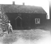 Anna Larsson (f. Hasselbergs) med dottern Berit. De står utanför Salves gård som var Annas morföräldrars bostad. 
Gården byggdes upp efter branden i Hällesåker år 1830.