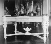 Ett ljust, snidat bord med tre urnor stående ovanpå. I bakgrunden ser man statyn 
