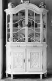 Vitrinskåp i ljus färg med ett par inglasade dörrar upptill och två luckor nertill. Skåpet innehåller porslinsföremål. Gunnebo slott 1930-tal.
