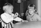 Två barn som sitter vid ett bord. Det vänstra barnet håller en äppleskrott i handen. Holtermanska daghemmet 1953.