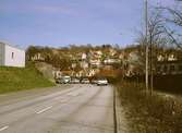 Kvarnbygatan österut, april 1993. Till vänster ligger parkeringslängor tillhörande bostadsområdet Silverskatten. Rakt fram ses villor i Mölndals Kvarnby och till höger muren som delar Kvarnbygatan och Papyrus Pappersindustri. Relaterat motiv: 2003_0327.