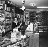 En kvinna står bakom disken i Åkerströms manufakturaffär på Sörgårdsgatan 2 i Krokslätt, okänt årtal. Relaterade motiv: 2003_0704 - 0708.