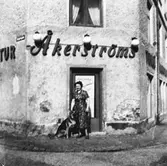 En kvinna och en schäfer står utanför Åkerströms manufakturaffär på Sörgårdsgatan 2 i Krokslätt. Okänt årtal.