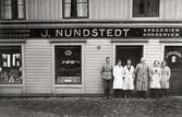 Personalen står utanför J. Nundstedts butik på Gamla torget i Kvarnbyn, 1930 - 1935. Från vänster: Åke Nundstedt (1913-1975), Majken Nundstedt (född 1911), ett biträde, Johannes Nundstedt (1878-1944), ett biträde, Vivi Nundstedt (född 1915). 
Sonen Åke Nundstedt fortsatte även senare som handlare, då i Göteborg. Relaterat motiv: 2004_0158.