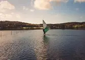 En person som vindsurfar över Berget 1:135, Lindome i oktober 1998. Lindomeån har svämmat över fälten mellan gården och gamla riksvägen (ganska vanligt). Och anledningen till att vindsurfaren är fotad är att det var märkligt att se en vindsurfare här. Relaterat motiv: 2004_0902.