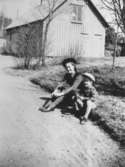 Rosa Pettersson (född Krantz) och en liten pojke sitter i gräset vid gårdsplanen till personalbostaden i Stetered, okänt årtal. En vedbod syns i bakgrunden.