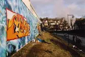 Graffiti längs Kvarnbygatan i Mölndal, år 2003. Till höger ses muren tillhörande Papyrusområdet och i bakgrunden skymtar Mölndals kvarnby. Relaterade motiv: 2007_0266 - 0284.