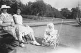 Sommar i Lindome, början av 1940-talet. Två kvinnor sitter på en bänk och ett litet barn sitter i en kärra. I bakgrunden ses tågspår.