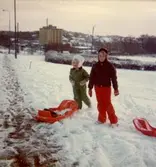 Christine och Ingela med pulkor vid Delbancogatan i Mölndal, december 1976.
Den gamla SOAB-cisternen i bakgrunden.