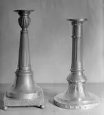 Två ljusstakar av metall med varierande höjd och design, ståendes på var sin sockel. Gunnebo slott 1930-tal.