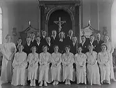 Interiör från koret i Varbergs kyrka, konfirmation 1935. Komminister Sven Gunnar Aronius (i mitten) med konfirmandgrupp. Från vänster, flickorna: Ulla Roswall, Gunnel Larzenius, Barbro Hylmö, Gullan Johansson från Stafsinge, Inga-Kerstin Svensson från Harplinge, Birgit Hallberg, Evy Antonsson från Tvååker, Gudrun Lauritzon, Lilli Svensson. Mellersta raden, pojkarna: Artur Lindberg, Alf Andersson-Ulmeborn från Okome, Lennart Bengtsson, Nils Liljegren, Karl-Erik Hansson, Filip Nilsson, Sven Wildheim, Erling Johansson. Översta raden: Per-Olof Johansson från Göteborg, Stig Svensson från Stafsinge, Lennart Olsson, Torsten Lennart Olsson från Stafsinge.
