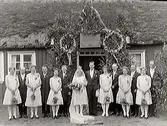 Brudparet Gustafsson (troligen) med brudfölje. Paret står under en äreport som rests över ingången till en låg stuga. Bruden har krona, och lång slöja och solglasögon(?) .  Brudens och tärnornas klänningar är knälånga, Herrarna bär frack.  Brudgumens namn är  troligen Gustaf Gustafsson. Brudens hamn okänt.