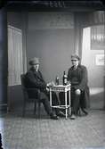 Två unga män i hatt vid ett bord med flaskor och glas, ateljébild.