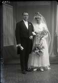Bröllopsbild. Ateljéfoto. Brudparet stående i helfigur. Bruden bär slöja och vacker krona, samt  håller stor bukett i vänster hand.